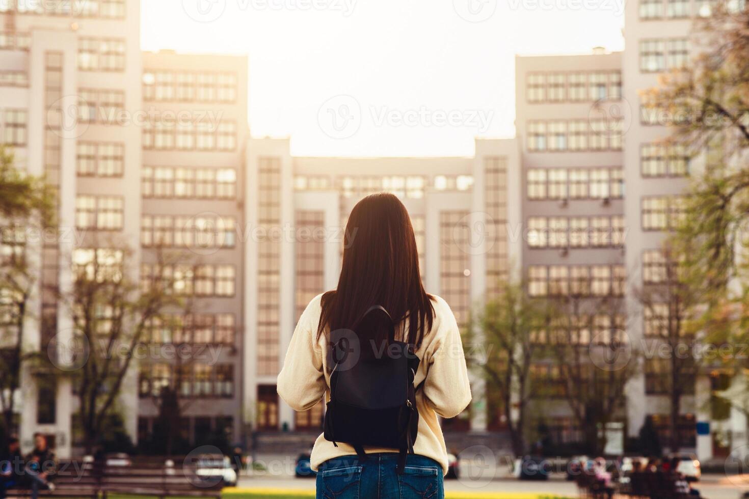 A woman stands in front of a towering building, looking up at its impressive height. The sleek architecture and glass facade of the building contrast against the sky, back view photo
