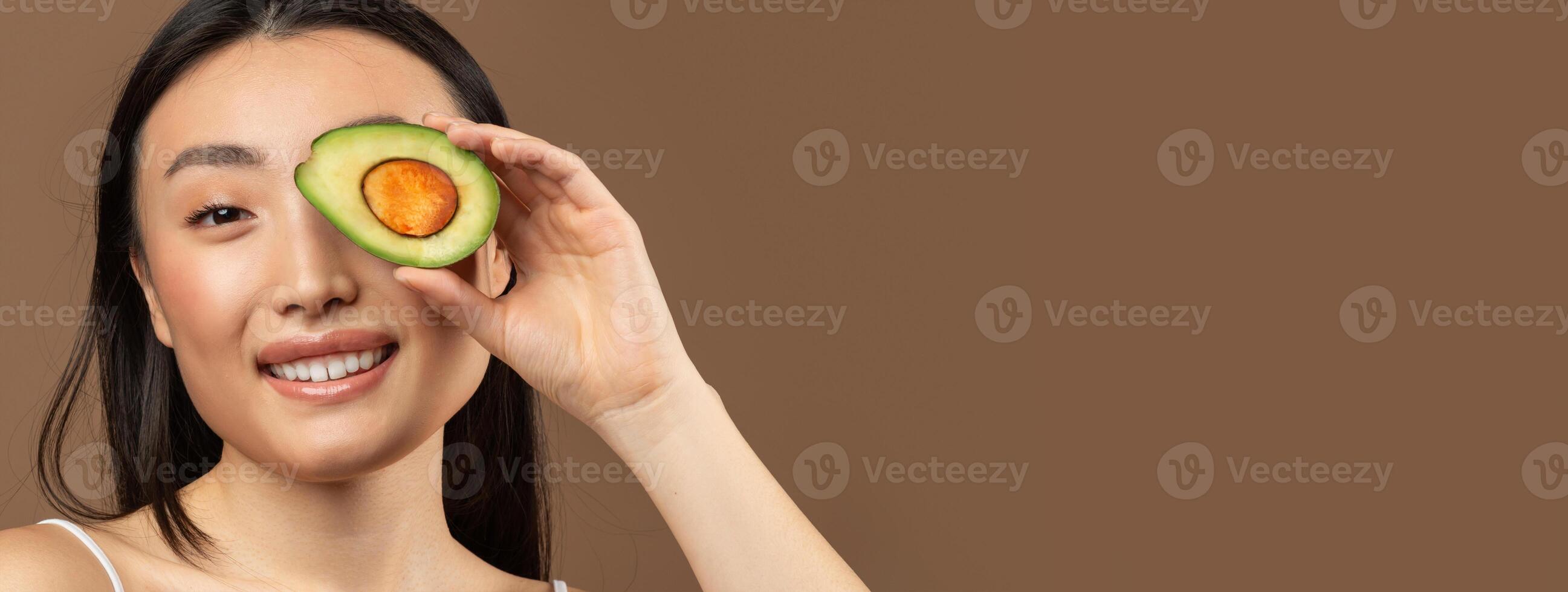 Portrait of young asian woman holding avocado half and covering her eye with it, smiling at camera isolated on brown studio background, panorama with copy space photo