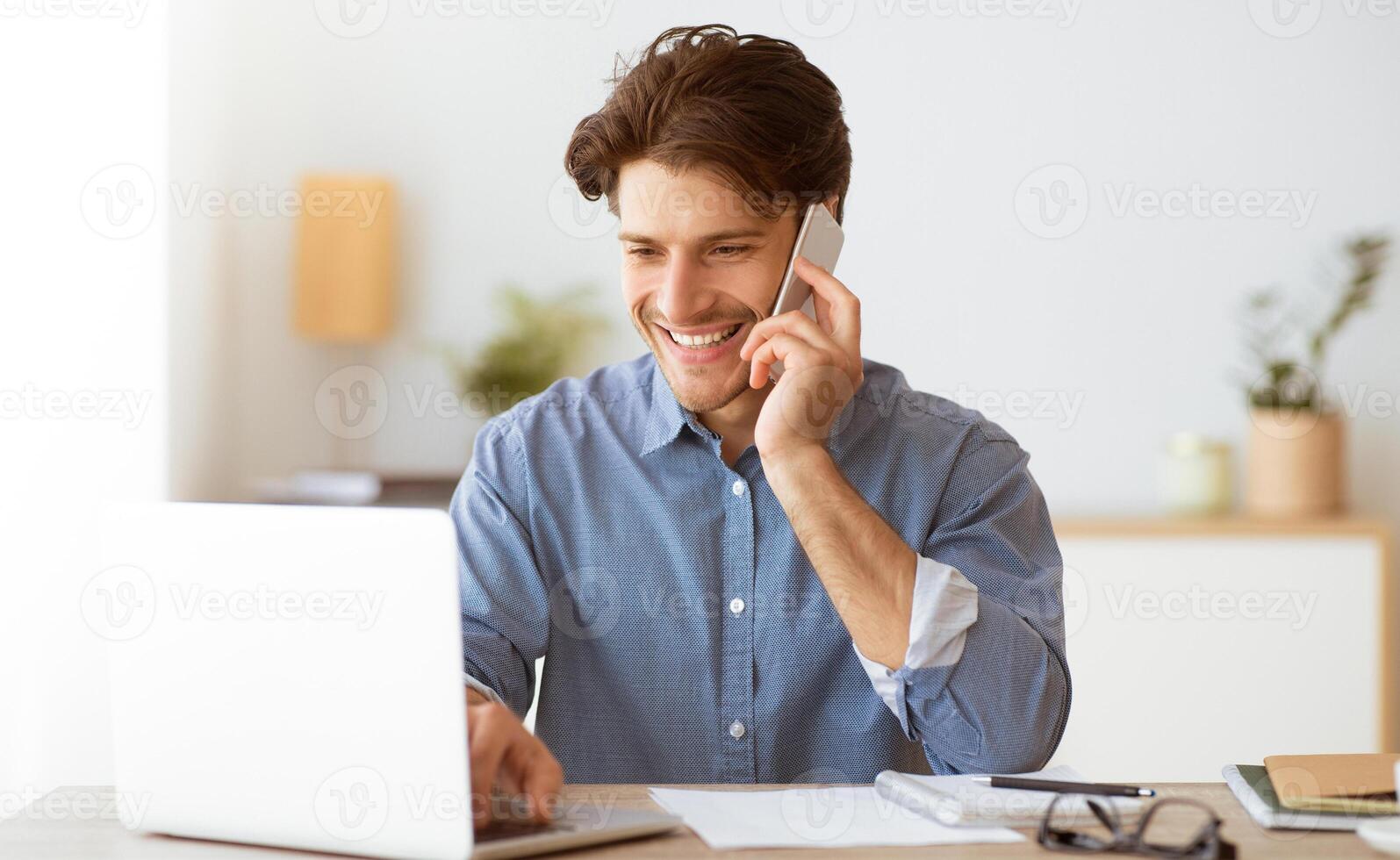 A man is smiling and talking on a phone while working on a laptop. He is seated at a desk and has paperwork in front of him. photo