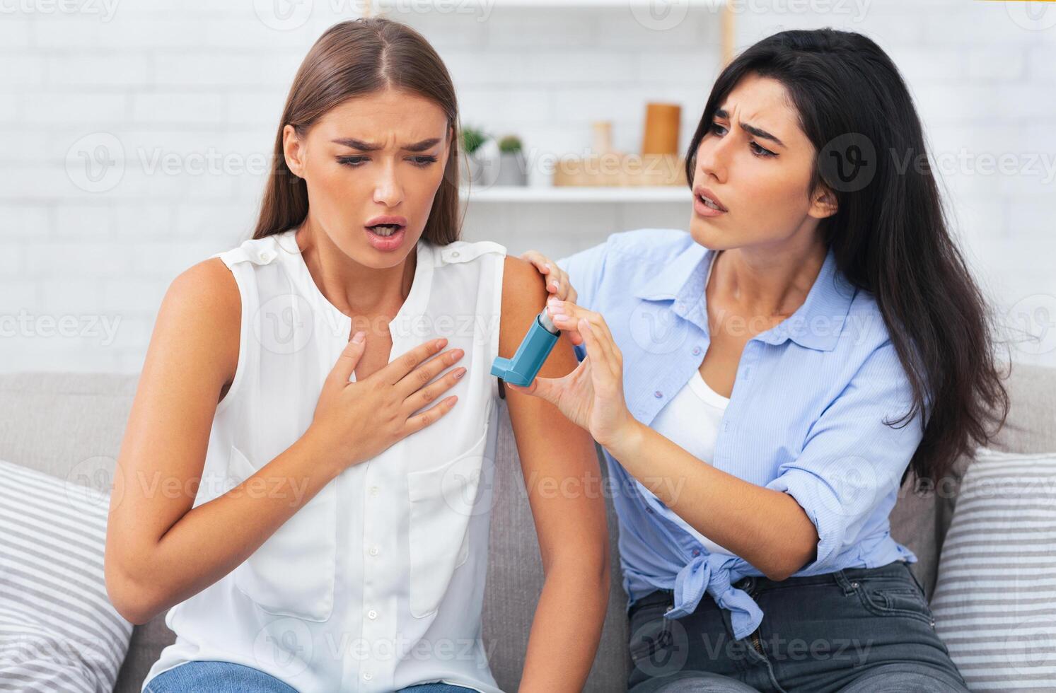 Girl Giving Asthma Inhaler To Friend Having Asthmatic Atack Or Respiratory Depression Sitting On Couch At Home. Selective Focus photo