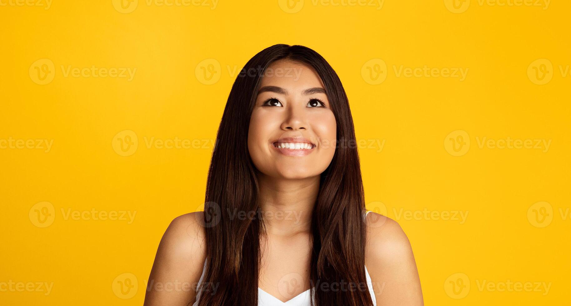 A young Asian woman with long dark hair is smiling as she looks up towards the sky. She is wearing a white tank top and standing in front of a solid yellow background. photo