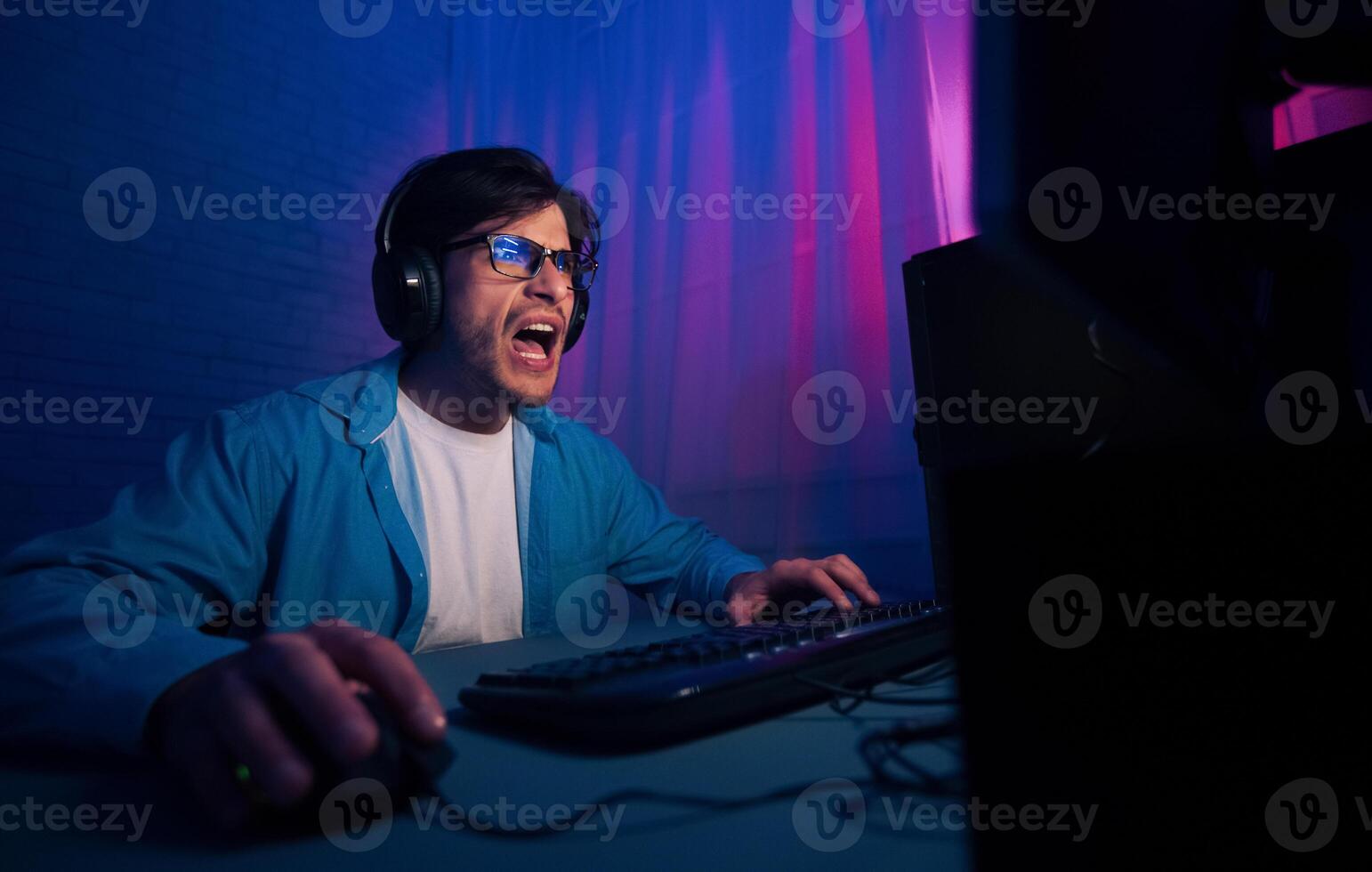 A man wearing headphones and a blue shirt sits in a dimly lit room, intensely focused on playing a game on his computer. The light from the monitor illuminates his face photo