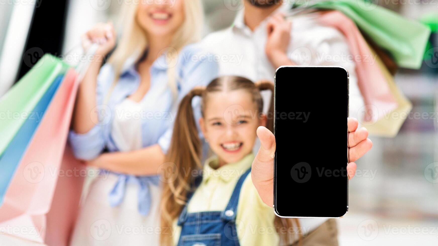 A smiling young girl holds a smartphone with a blank screen towards the camera. Her parents are blurred in the background, holding shopping bags. photo