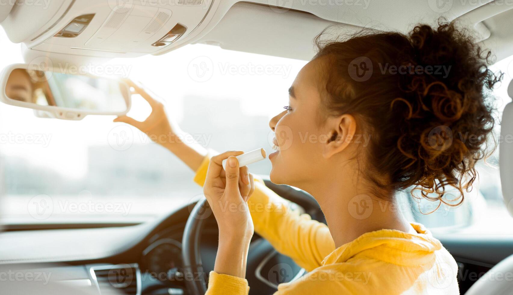 African American young woman with curly hair is sitting in the drivers seat of a car. She is applying lip balm to her lips while looking in the rearview mirror. photo