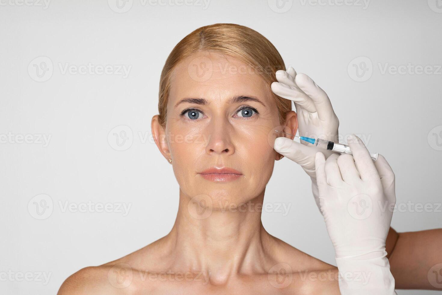 This image shows a woman with blonde hair receiving an injection to her cheek. She is looking directly at the camera, and a gloved hand is holding the syringe near her face. photo
