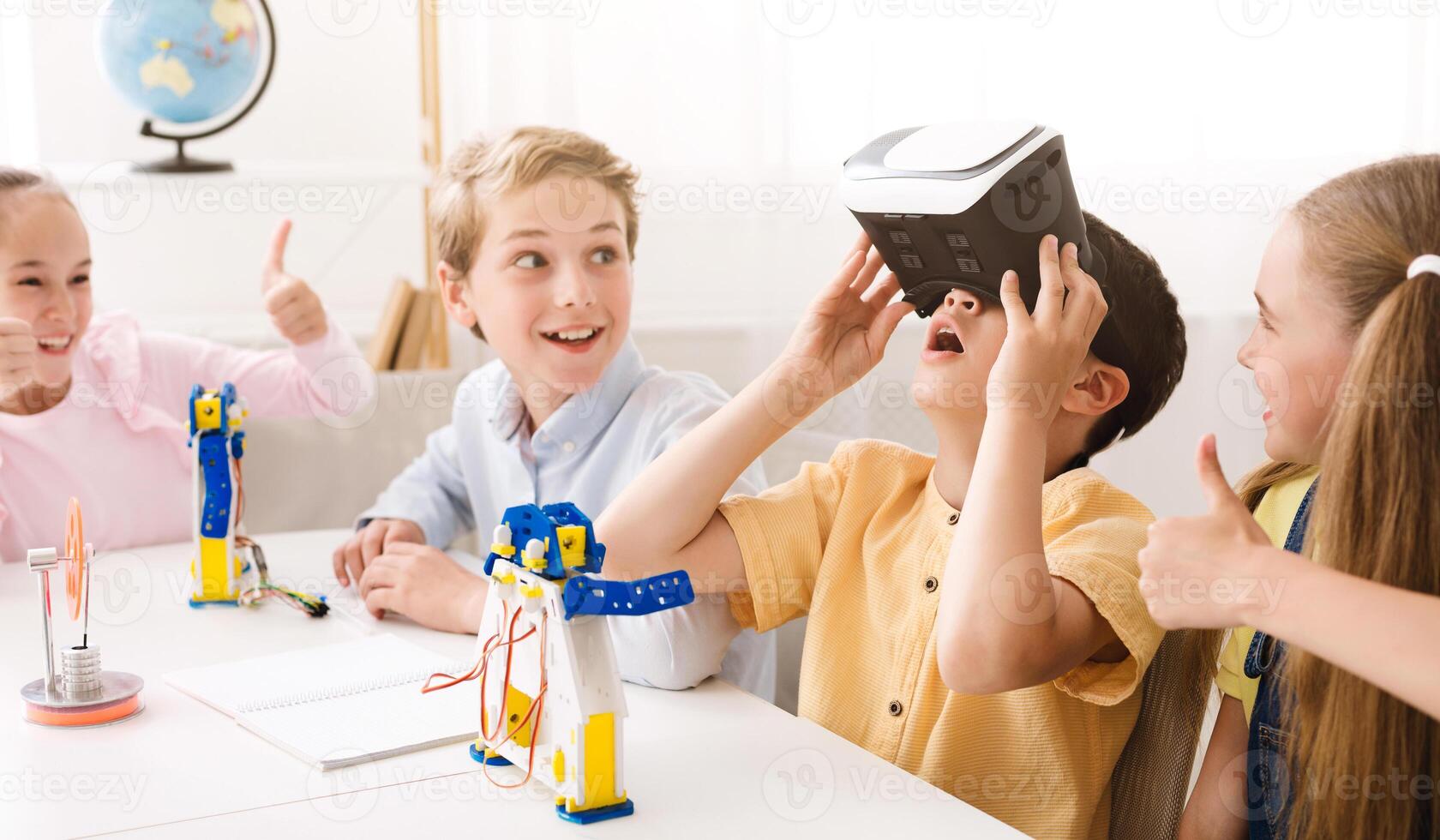 A group of children in a classroom are working on a project. One of the children is wearing a virtual reality headset while the others look on. They are all smiling and having fun. photo