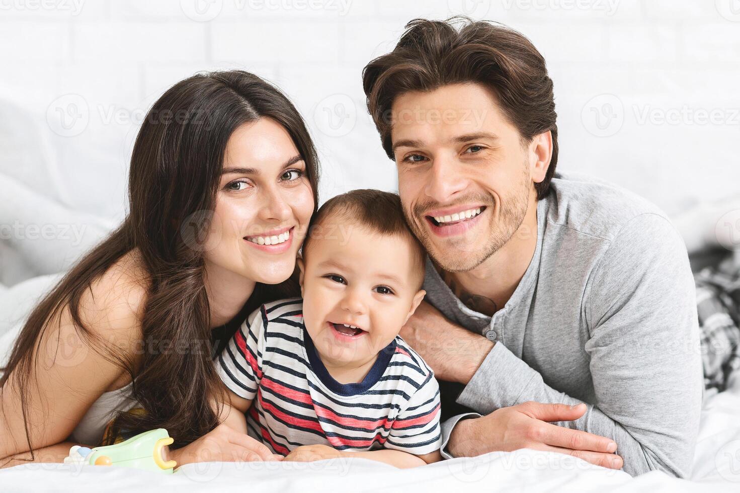 This image depicts a happy family of three - a mother, father, and their young baby - lying on a white bed together. The parents are smiling and looking at the camera photo