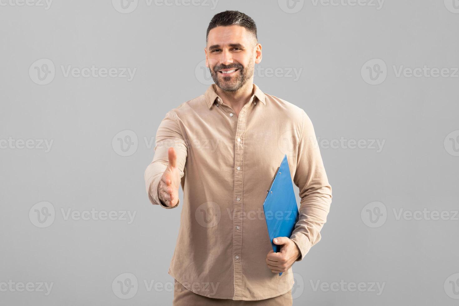 A well-dressed man smiles warmly as he extends his hand for a handshake. He holds a blue folder in his other hand, which may contain important documents. The background is plain and grey photo