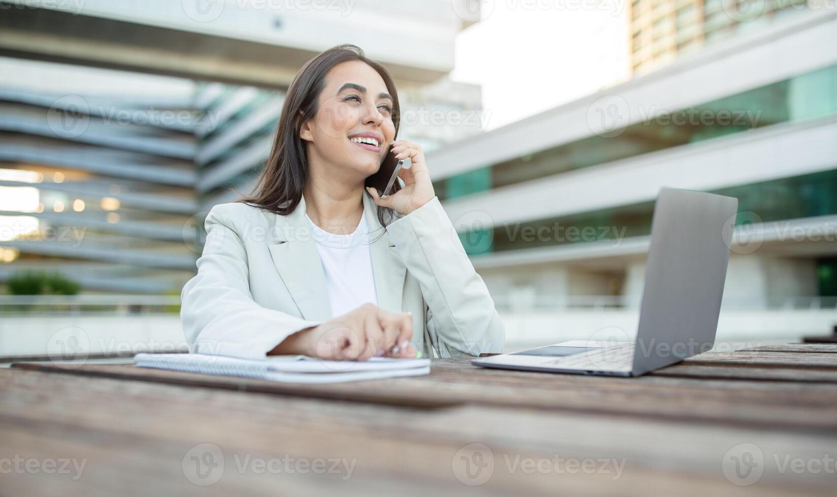 A young businesswoman sits outside, working on her laptop and taking a phone call. She is smiling and appears to be enjoying her work. The background features an office building with large windows. photo