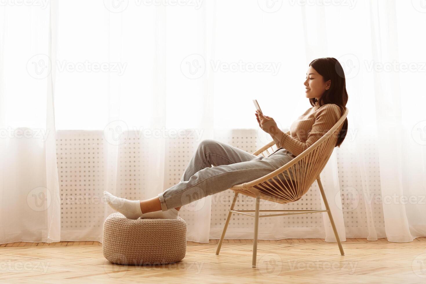 A woman is sitting in a woven chair and looking at her phone. Her feet are resting on an ottoman. photo