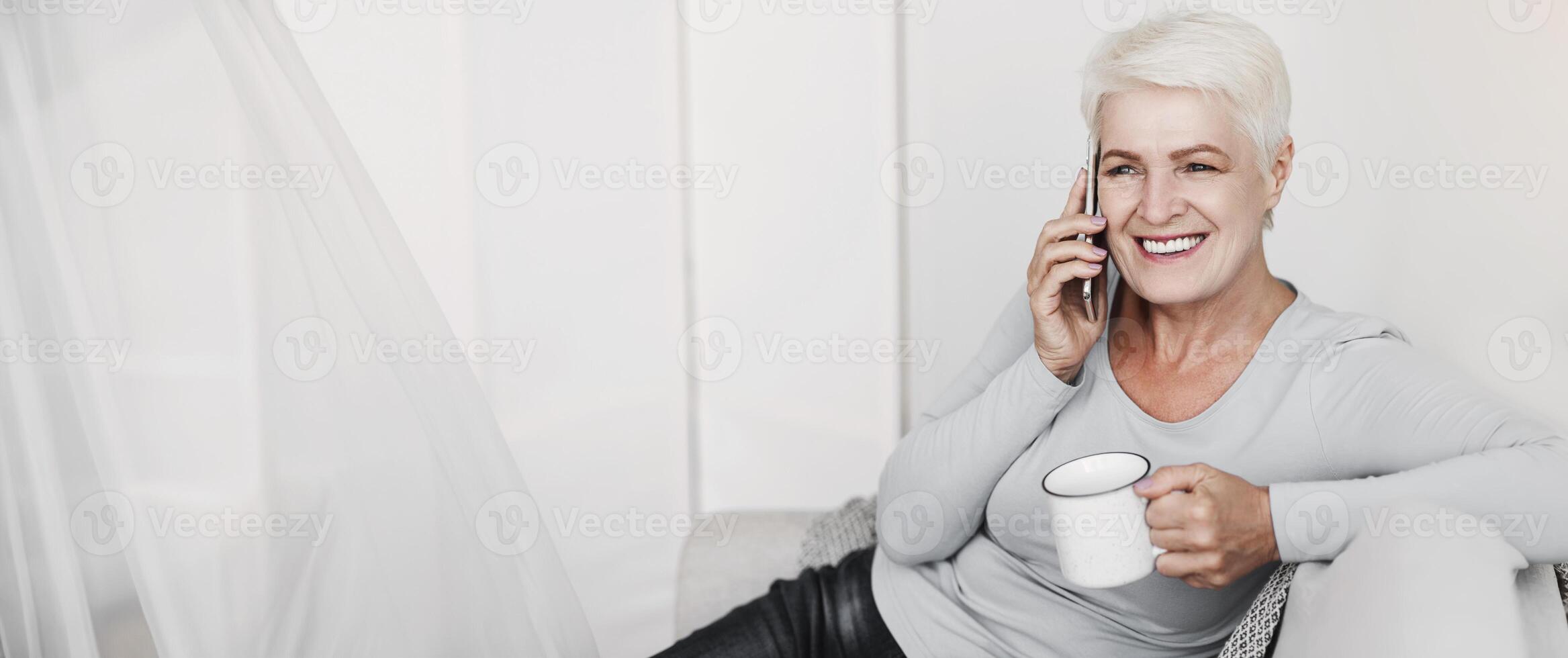 Smiling senior business woman drinking coffee and talking on smartphone, sitting at sofa at home, copy space photo