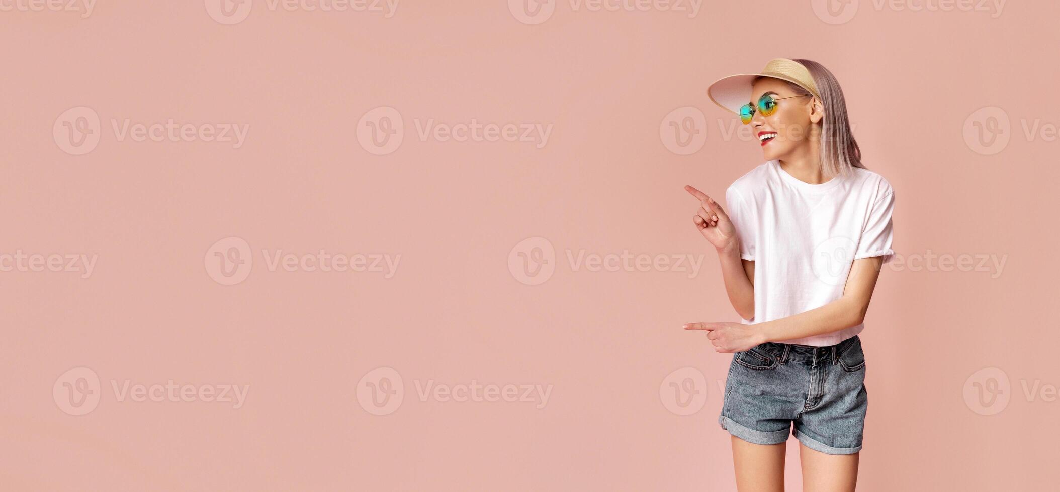 A woman in sunglasses and a hat is standing in front of a pink background. She is smiling and pointing to the left with her right hand, copy space photo