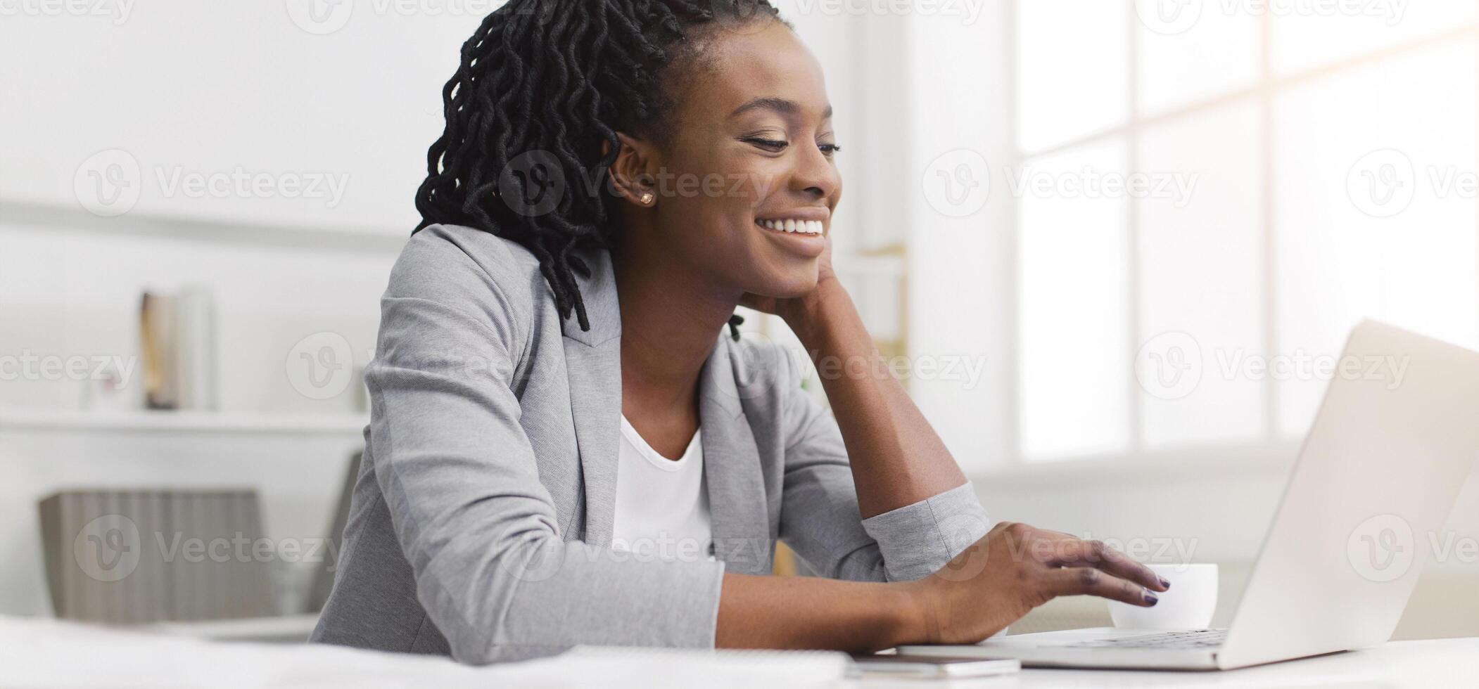 A hardworking African American lady is using her laptop in an office. The mood is productive and professional, with a tidy desk and modern decor, reflecting dedication to her work. photo