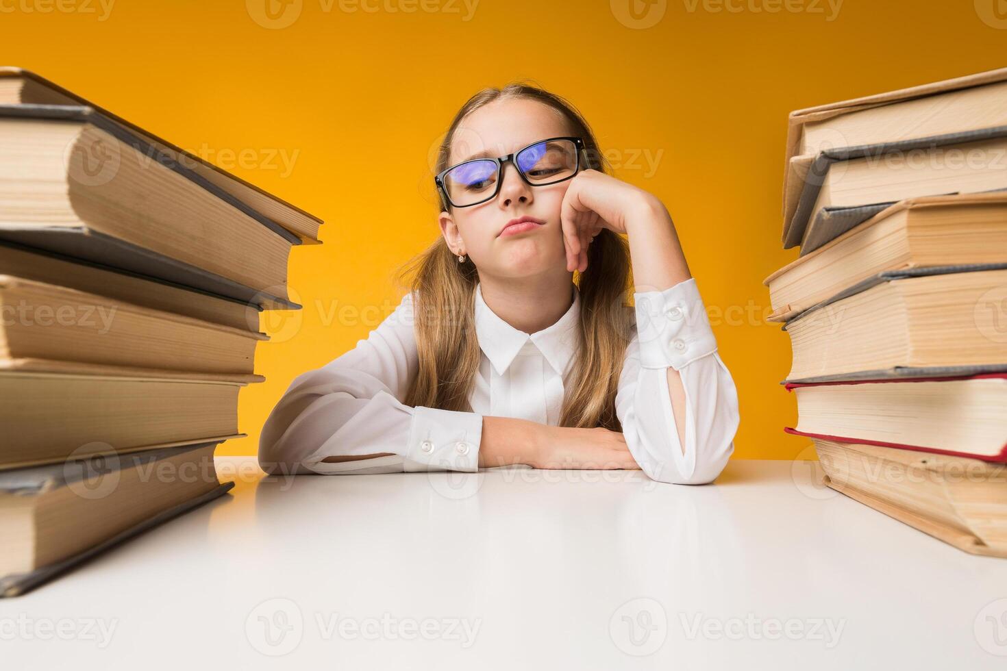 Boring Lessons. Sad Primary School Girl Sitting Between Two Book Stacks On Yellow Background. Studio Shot photo
