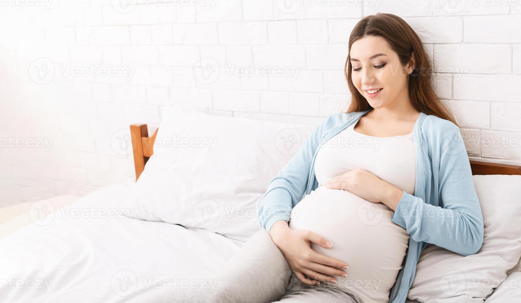 A pregnant woman is seated on a bed. She is resting comfortably, with her hands gently cradling her baby bump. The room is softly lit, creating a peaceful atmosphere. photo