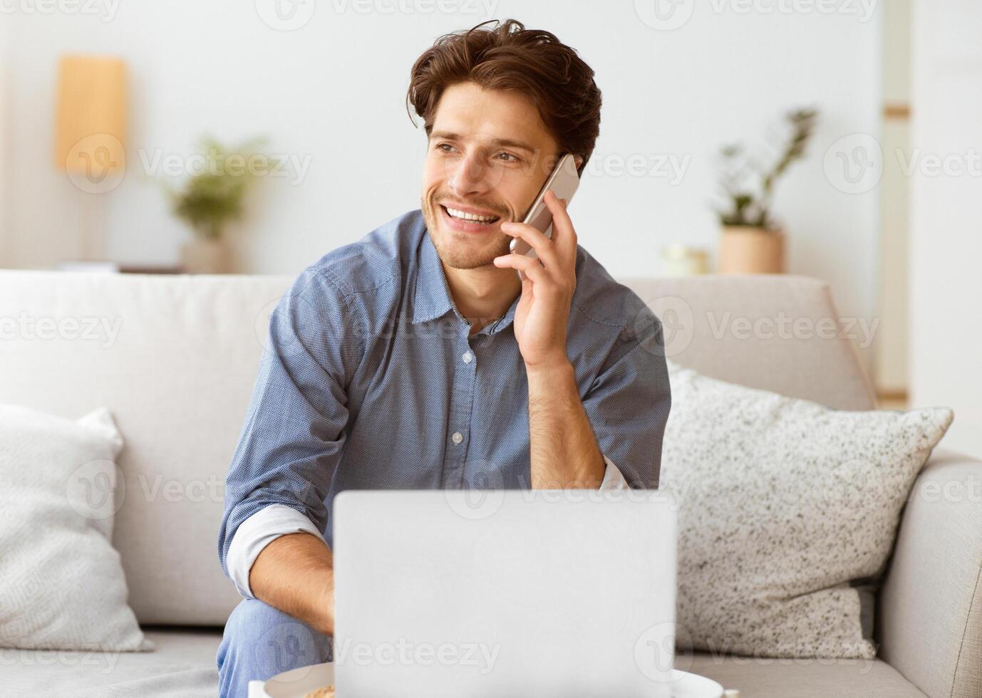 A man smiles while talking on his phone and using his laptop, sitting on a sofa in his home. photo