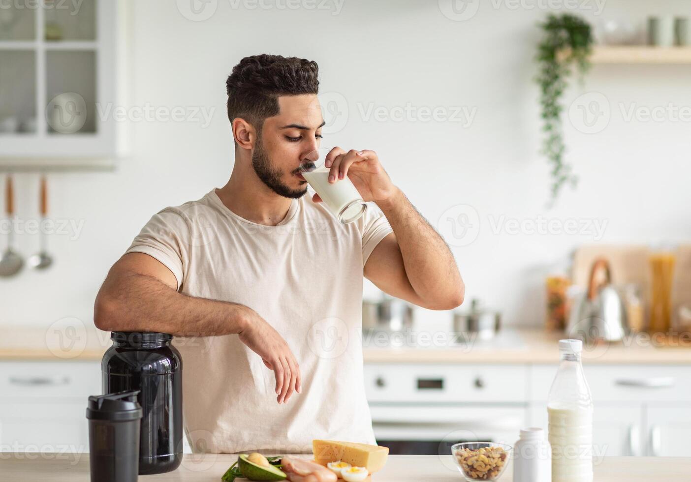 Healthy nutrition for muscle gain and weight loss concept. Athletic young Arab man drinking protein shake or milk, standing near table with healthy wholesome products at kitchen, copy space photo