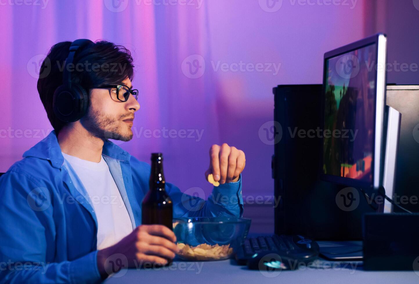 This image shows a young man playing games while wearing headphones. He is sitting in front of a computer with a controller in his hand. He is enjoying snacks and a beer while he plays. photo