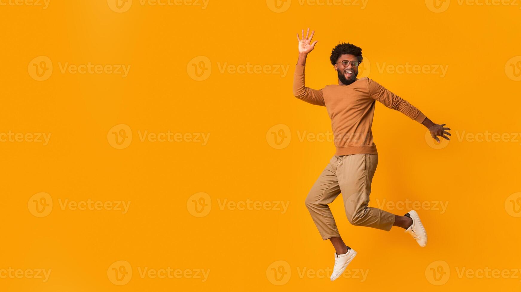 Cheerful black guy in glasses jumping over orange background photo