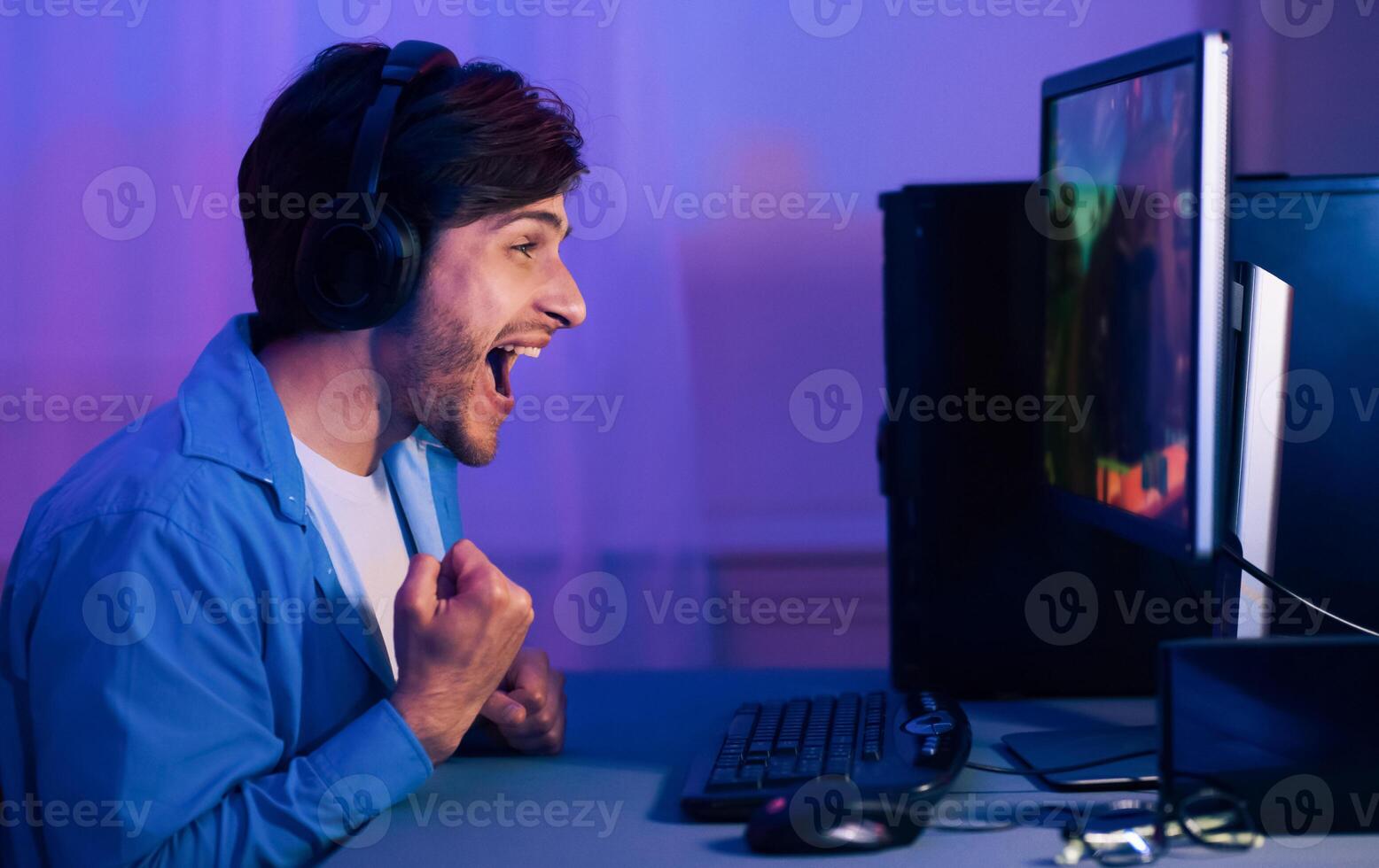 A young man with headphones on sits at his computer desk and celebrates a victory in a game. His mouth is open in excitement and his fists are clenched. photo