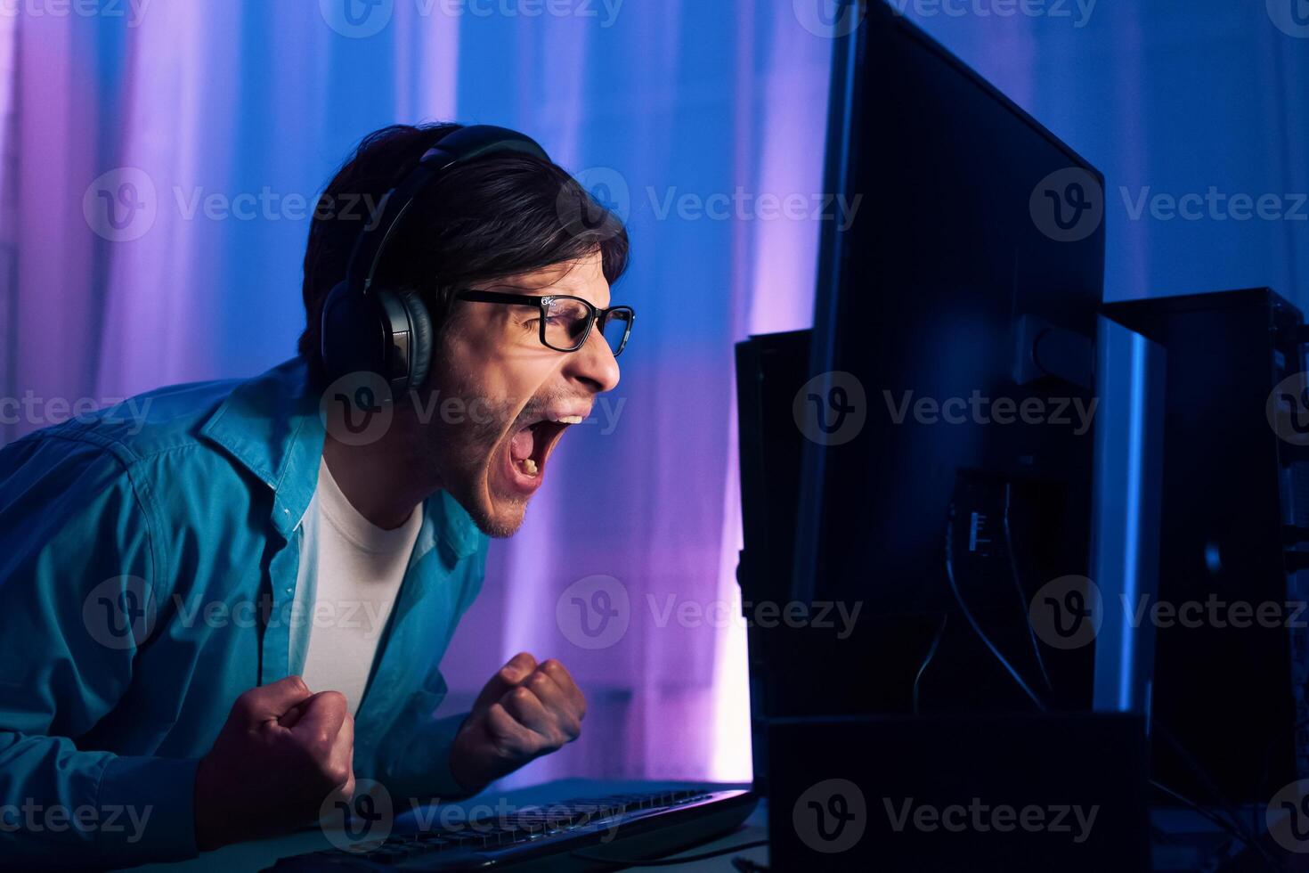 A young man wearing headphones and glasses sits at his computer and screams with excitement while playing a game. The room is dimly lit, and the monitor is brightly lit photo