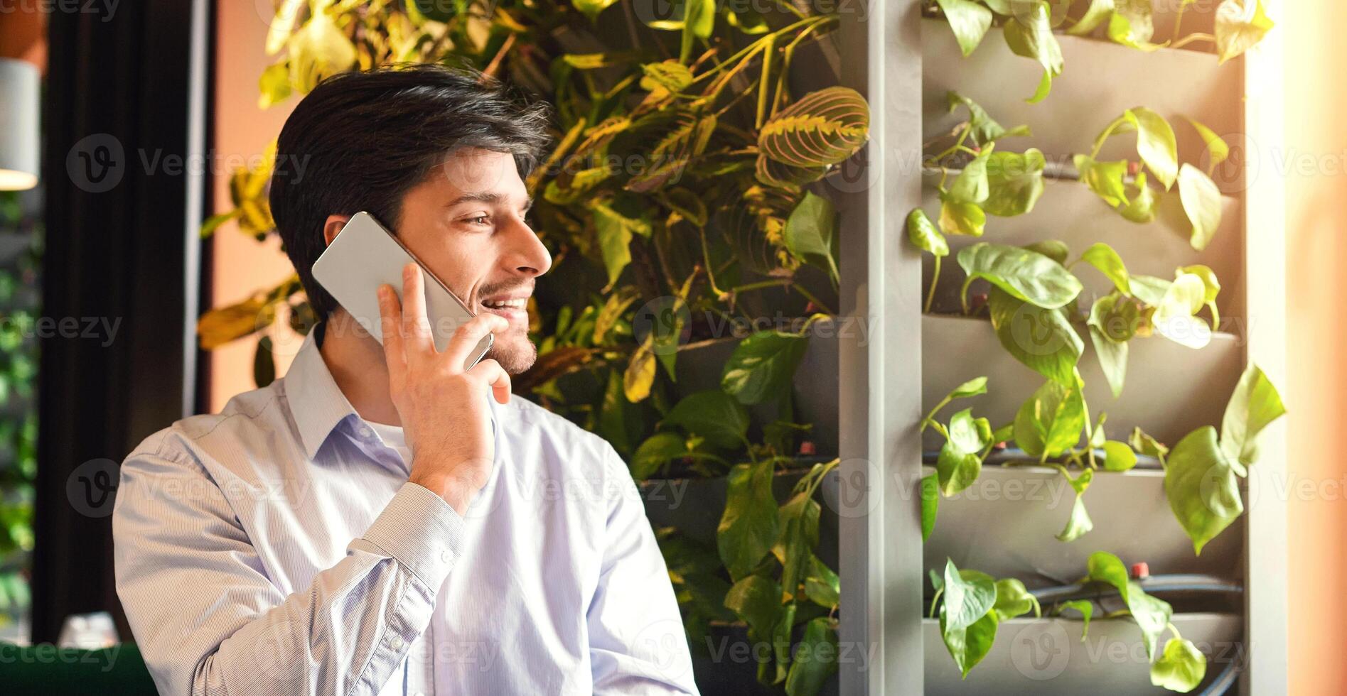 Handsome man talking on phone and smiling at modern cafe, looking at empty space, sun flare, panorama photo