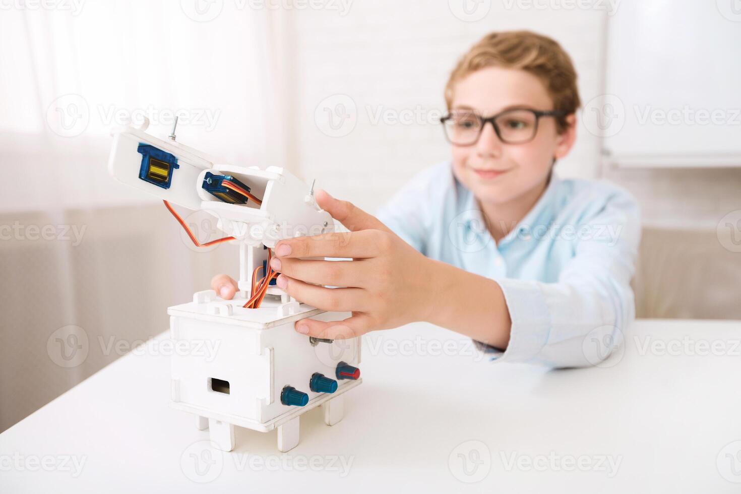 A young boy wearing glasses is seen at a desk, focused on assembling a white robotic arm model. He carefully aligns the components, demonstrating his interest in robotics and STEM learning. photo