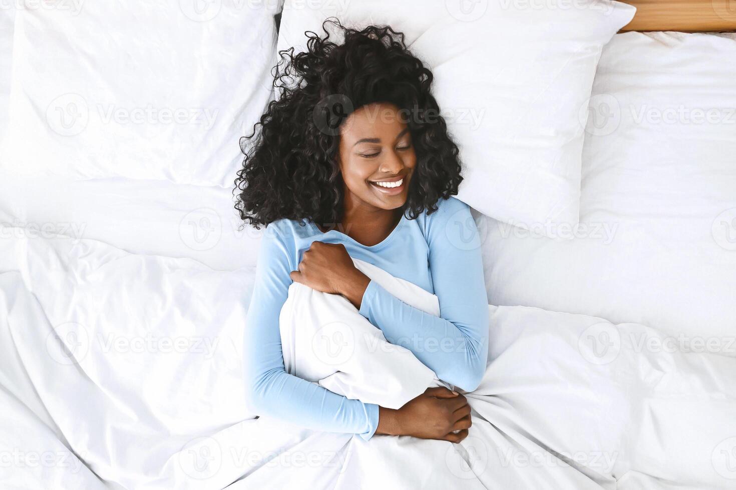 Young well-slept african woman waking up in bed, smiling and holding blanket with closed eyes, empty space, top view photo