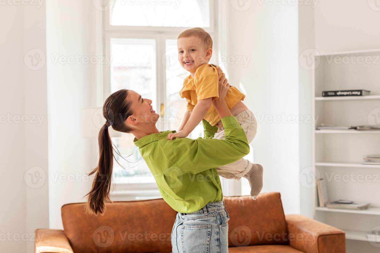 A woman cheerfully holds a child up in the air, both smiling. The child arms are outstretched, enjoying the playful moment. Mother face shows joy and love for her son photo