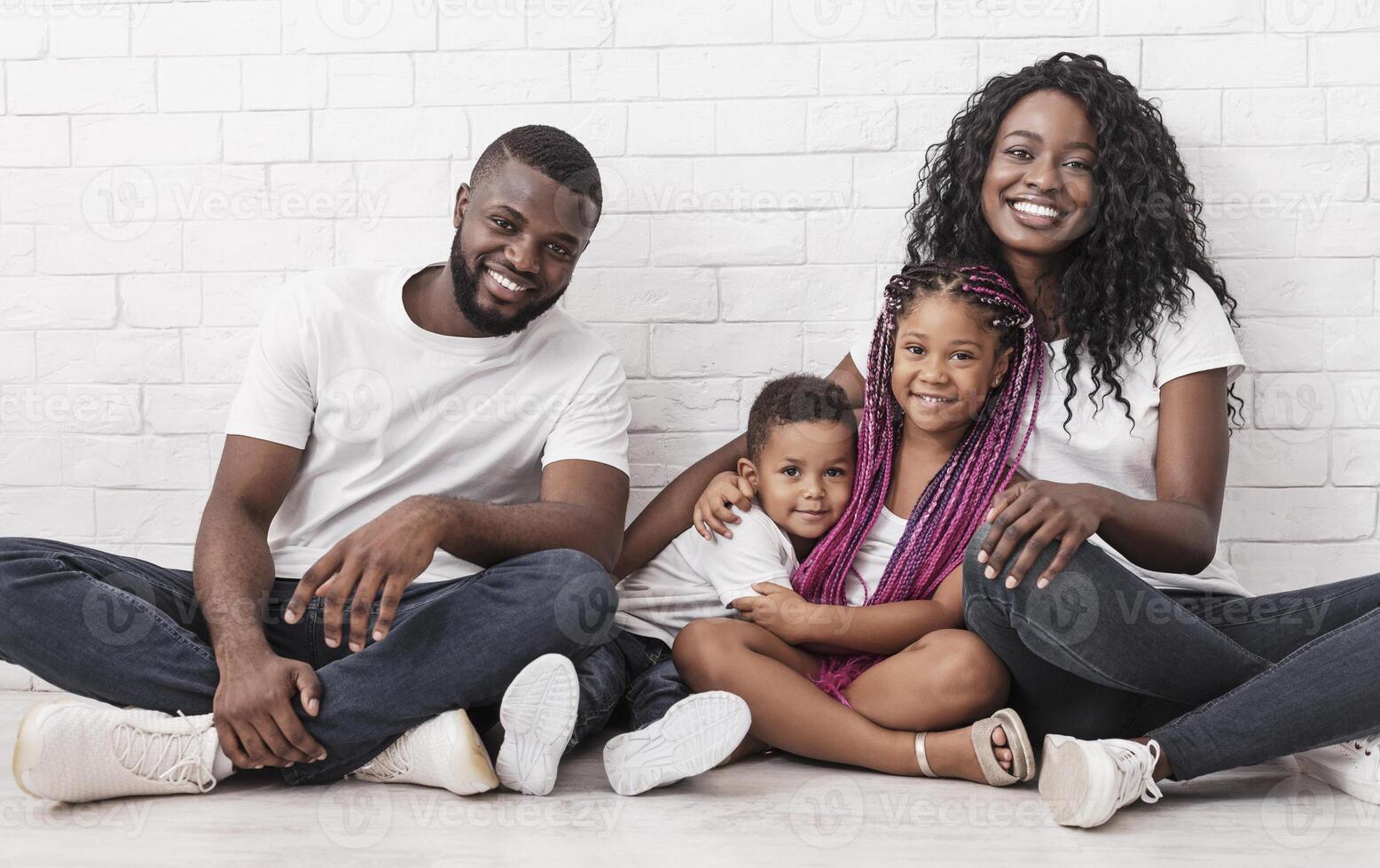 Joyful afro family mother, father and two little children sitting on floor and cuddling over white brick wall background. photo