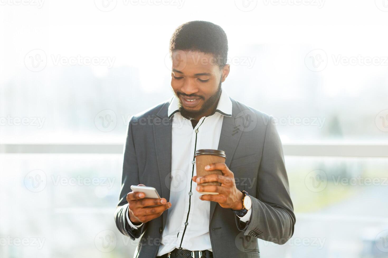 Smiling black businessman holding a coffee cup while checking his smartphone in a bright, modern setting. photo