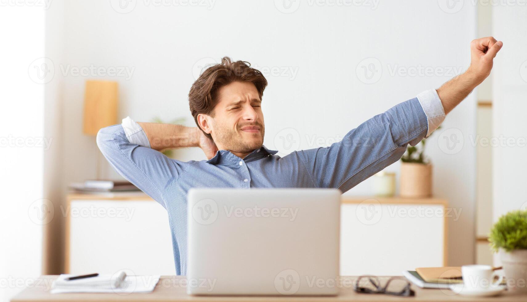 A man sits at a desk in his home office, stretching his arms and back. He is looking slightly tired. photo