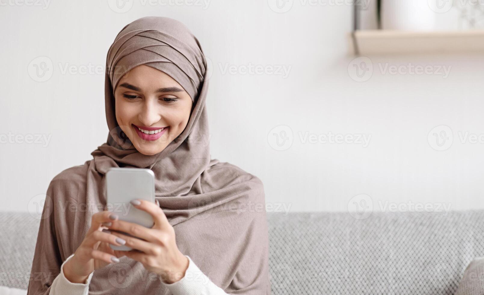 Portrait of millennial muslim girl in hijab using smartphone at home, messaging or browsing social networks, sitting on sofa in living room photo
