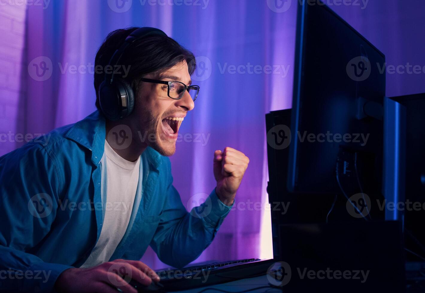 A young man wearing headphones and glasses is seen celebrating a victory while playing a game on his computer at night. The image captures the intensity and excitement of competitive gaming photo