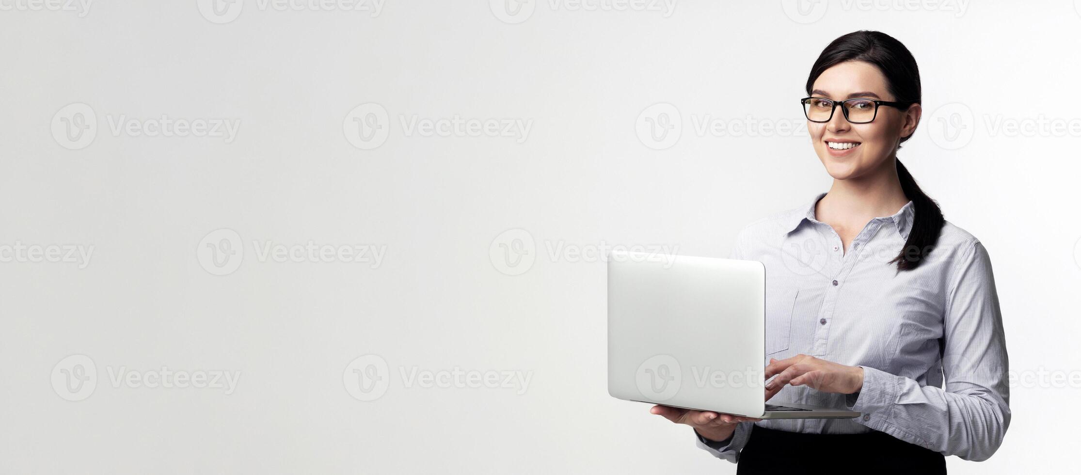 A smiling businesswoman in a white shirt holds a laptop in front of a white background. photo