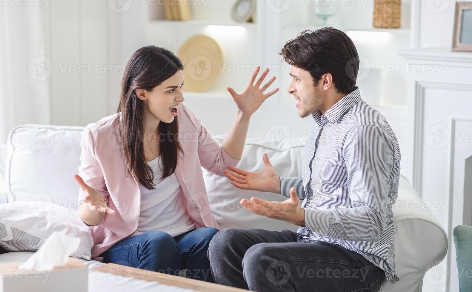 A man and woman are engaged in a heated argument on a couch in a living room. They appear frustrated and are gesturing emphatically as they speak. photo