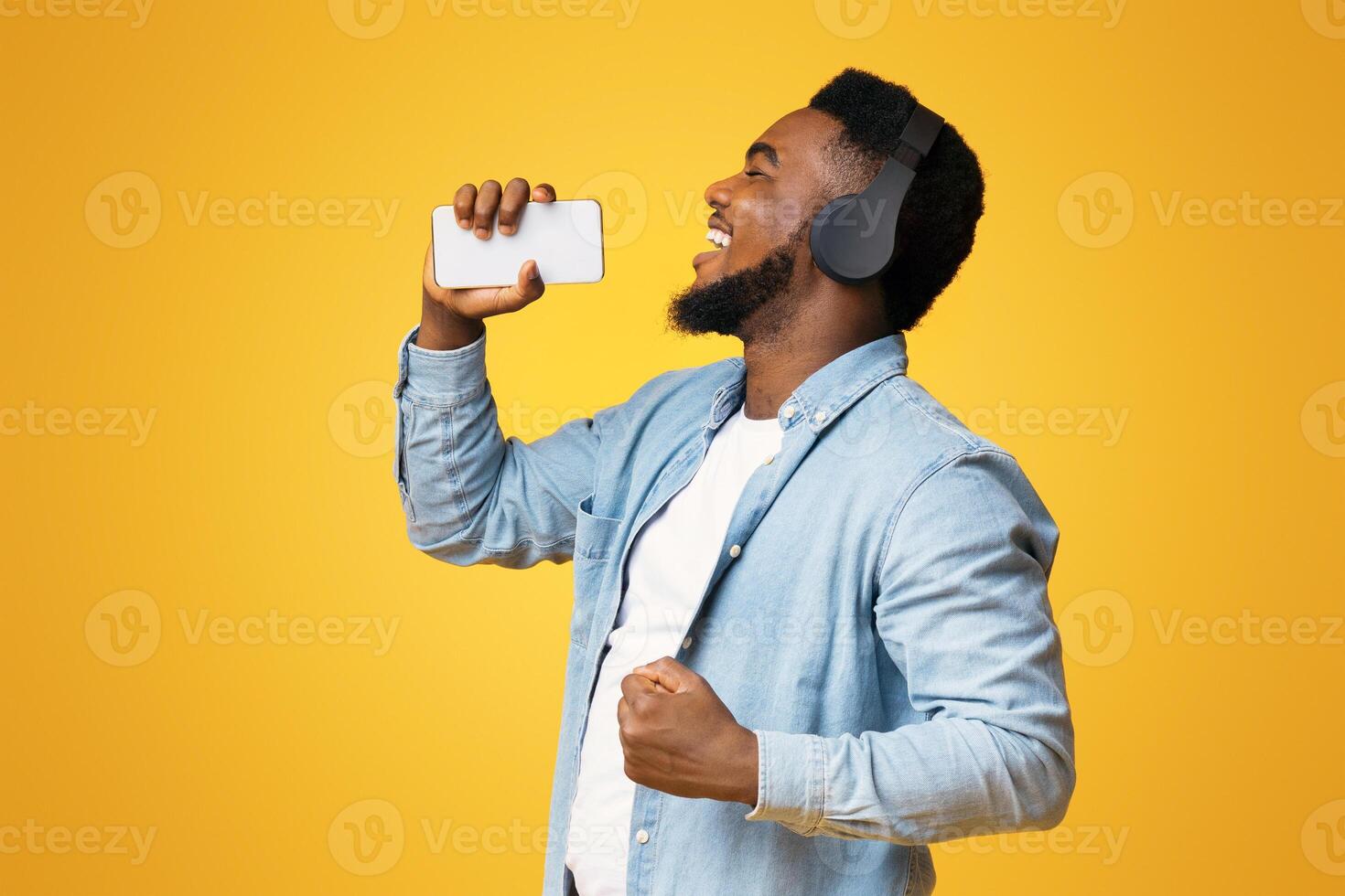 Reveal your talents. Cheerful african american man singing into smartphone like microphone on yellow background, side view photo