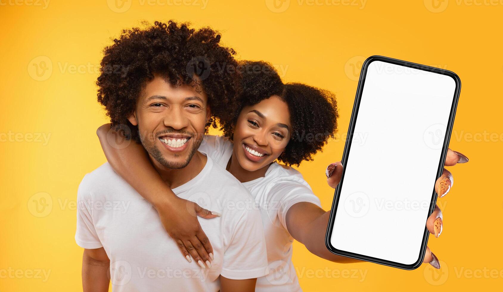 African American couple, a man and a woman, are smiling and looking at the camera. The woman is holding a smartphone with a blank screen in front of her. photo