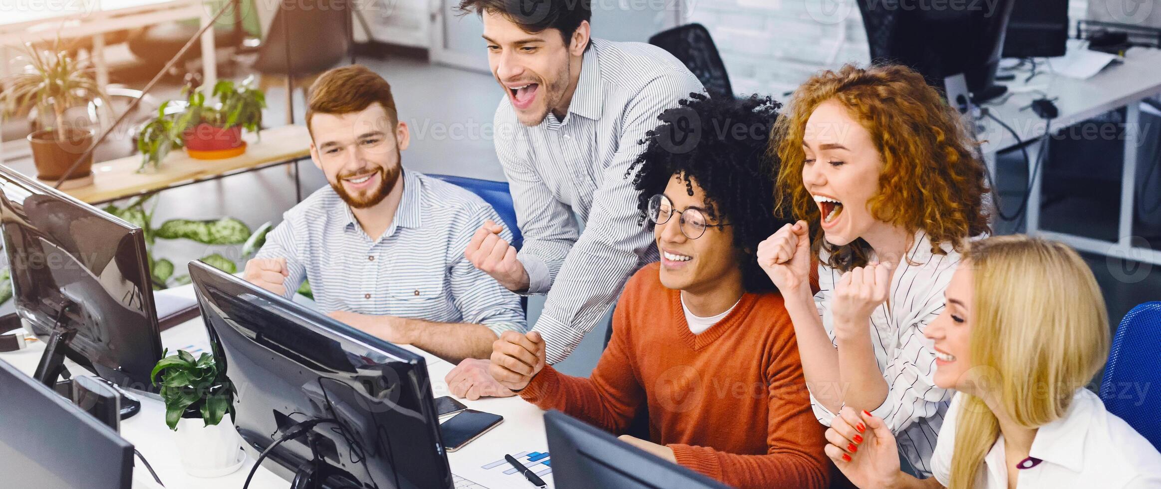 Young team of excited stock exchange traders celebrating success in office, looking at computer and shouying, panorama photo
