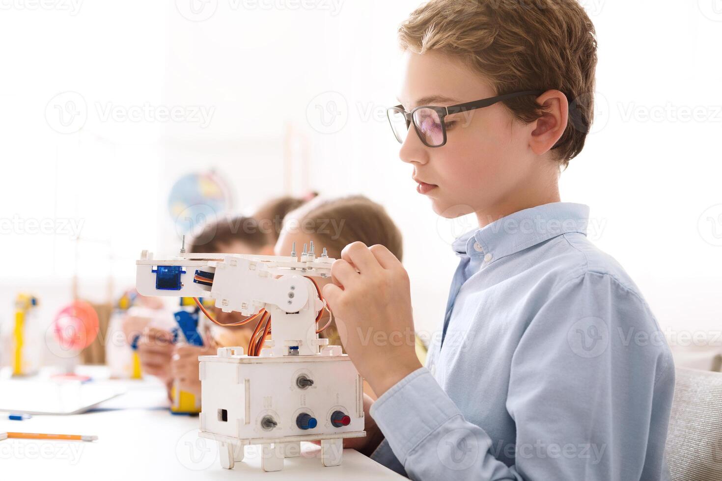 A young boy with glasses is sitting at a table in a classroom setting and is working on building a small, white robotic arm. He is holding a small piece of the robot in his hand. photo