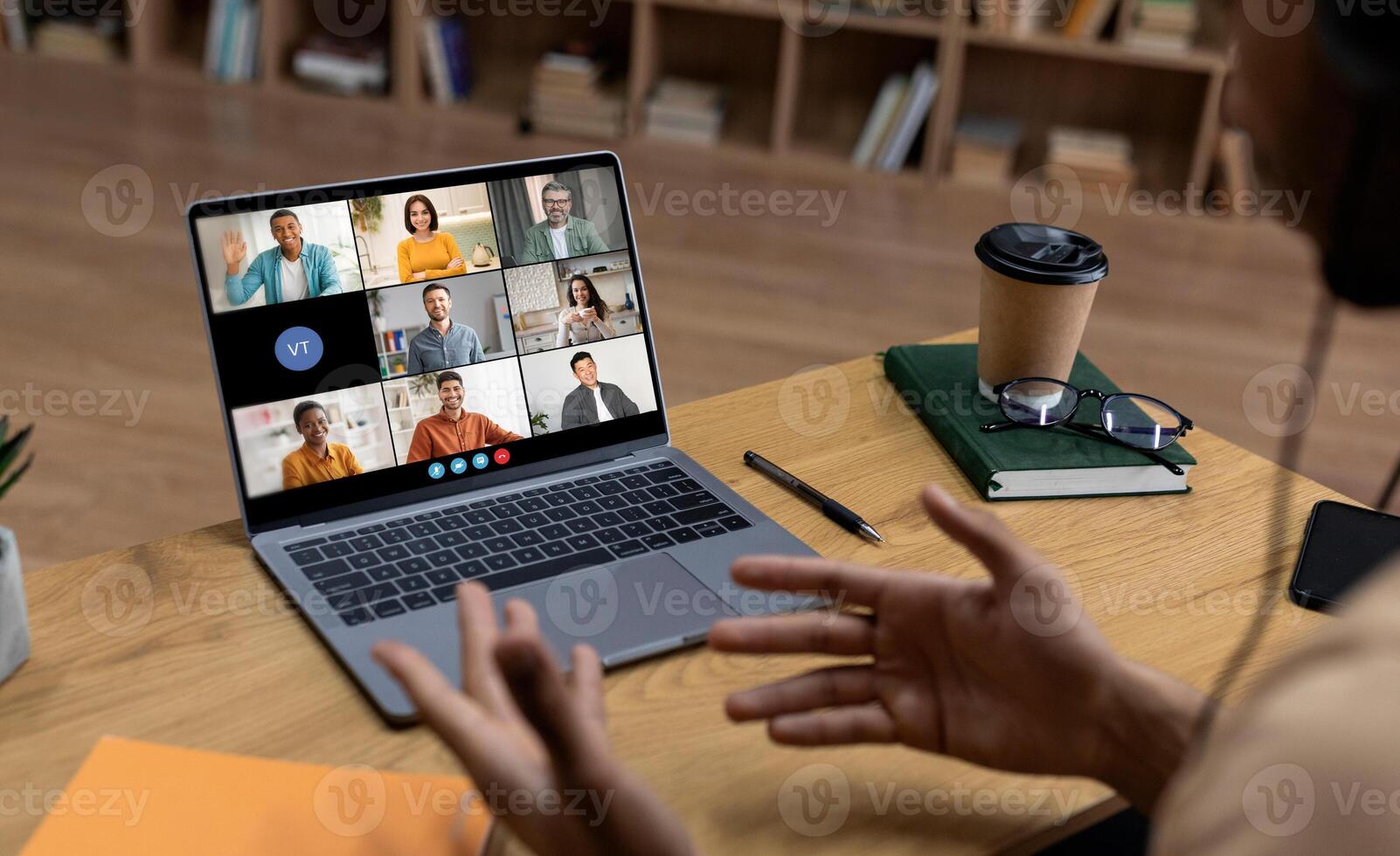 A person is sitting at a desk in front of a laptop computer, participating in a conference call. The laptop screen displays nine people in separate squares. photo