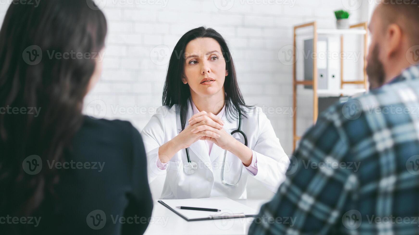 Lady doctor is sitting at a desk, consulting with a couple in a medical office. She is wearing a white coat and has a stethoscope around her neck. The couple is sitting across from her photo