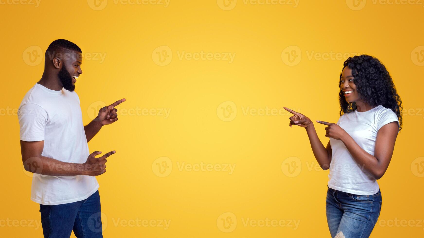 Joyful black couple smiling and cheerfully pointing away at empty space, yellow background photo
