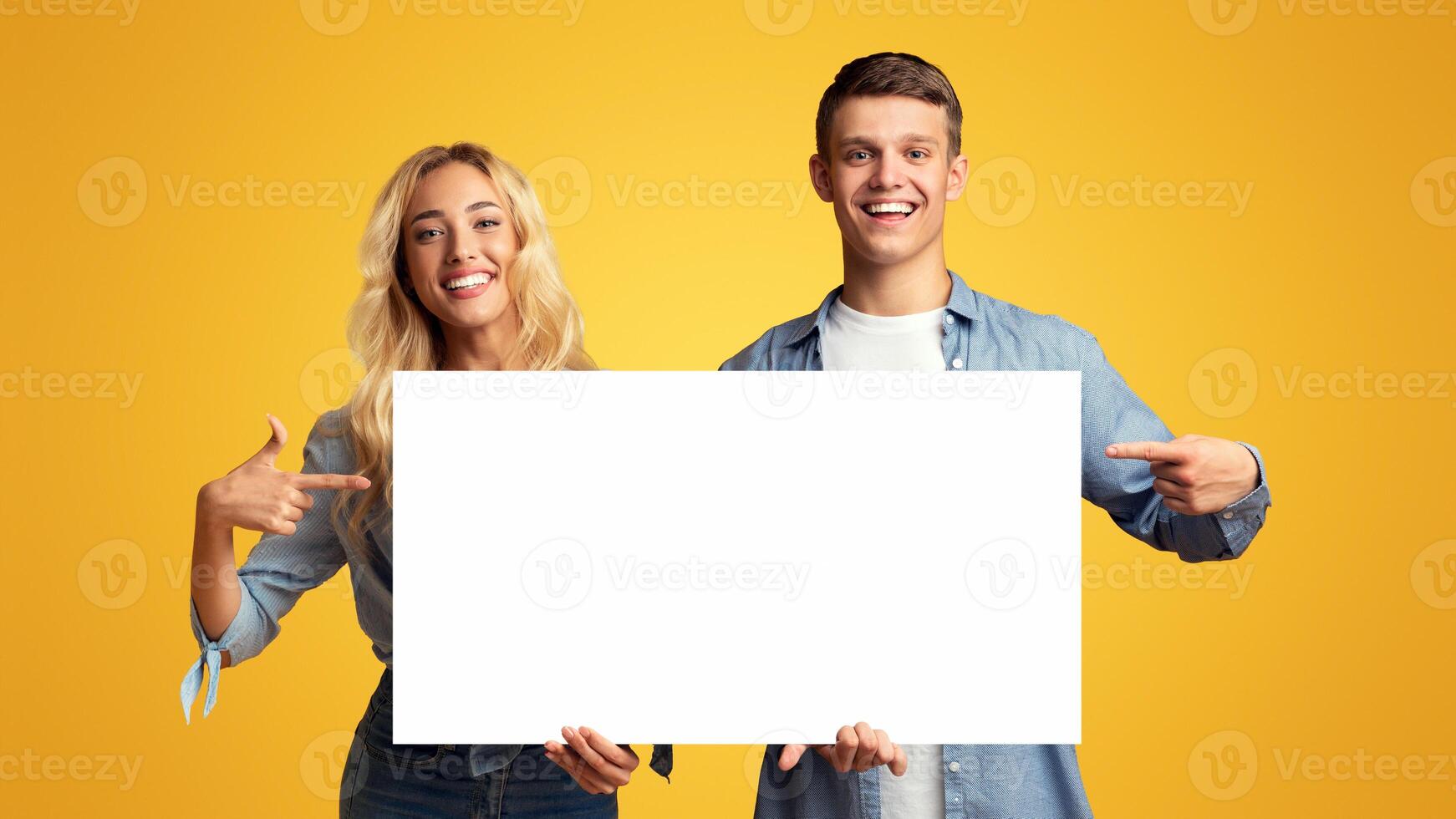 Place for your ad. Happy millennial guy and girl holding blank advertising board, pointing on it and smiling at camera over yellow background photo