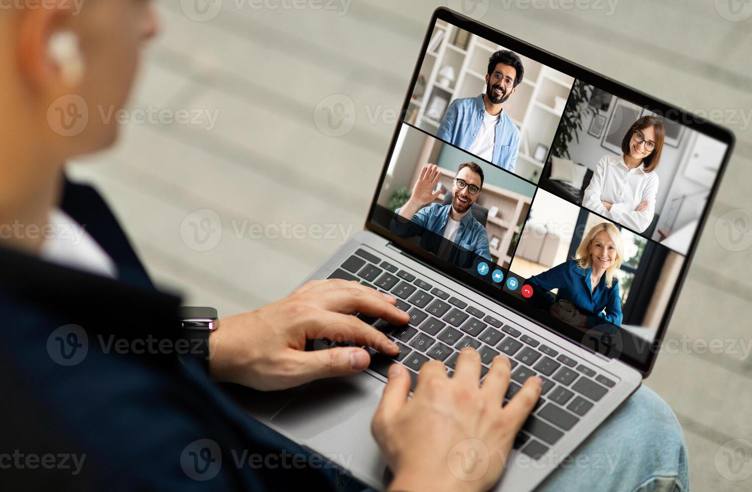 A man sits at a desk and participates in a conference meeting with four other people on his laptop. He is wearing a blue shirt and has his hand on the keyboard. photo