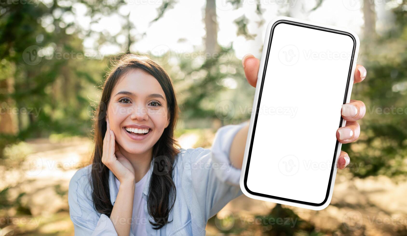 A young woman with long dark hair is smiling as she holds out a white smartphone with a blank screen, wearing a light blue shirt and is standing in a park with green trees and a blurred background. photo