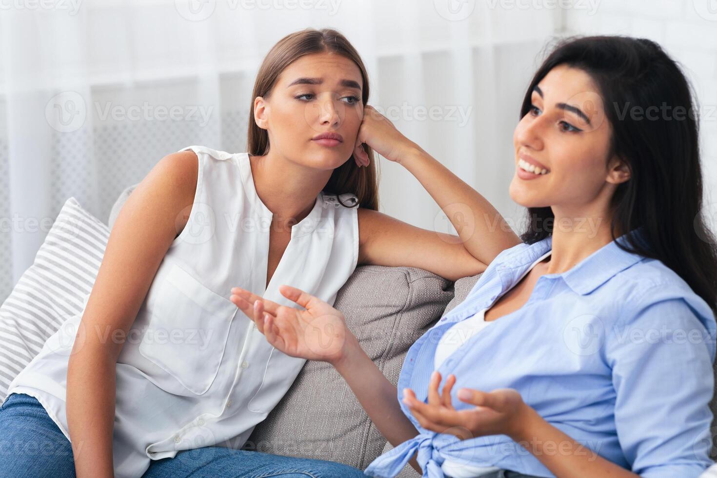 Jealous Woman Listening With Envy To Her Friend Bragging About Great Life Sitting On Sofa Indoor. Selective Focus photo