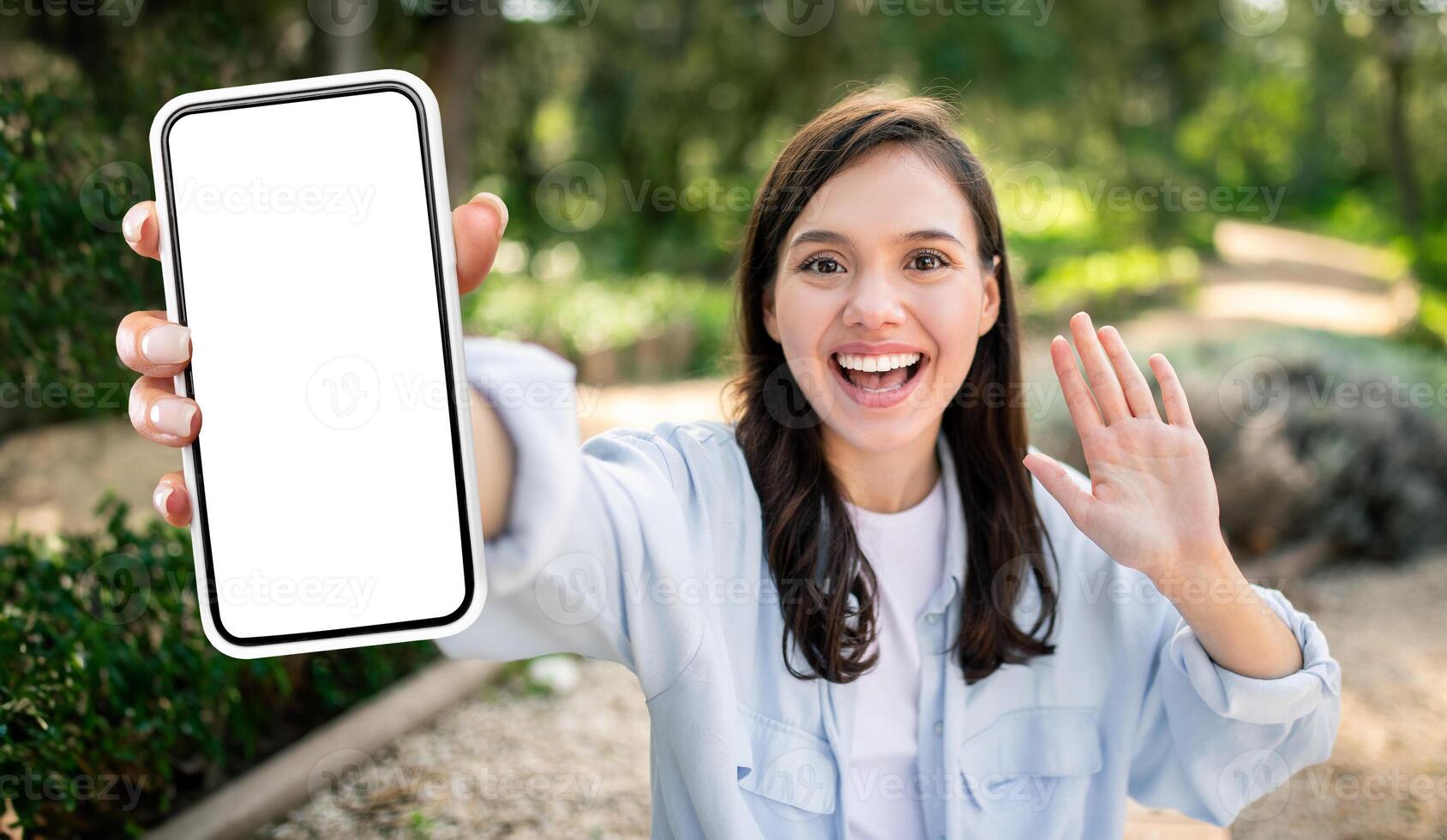 A young woman in a light blue shirt is outdoors, holding a smartphone with a blank screen in her right hand. She is smiling and waving with her left hand, as if she is on a call photo