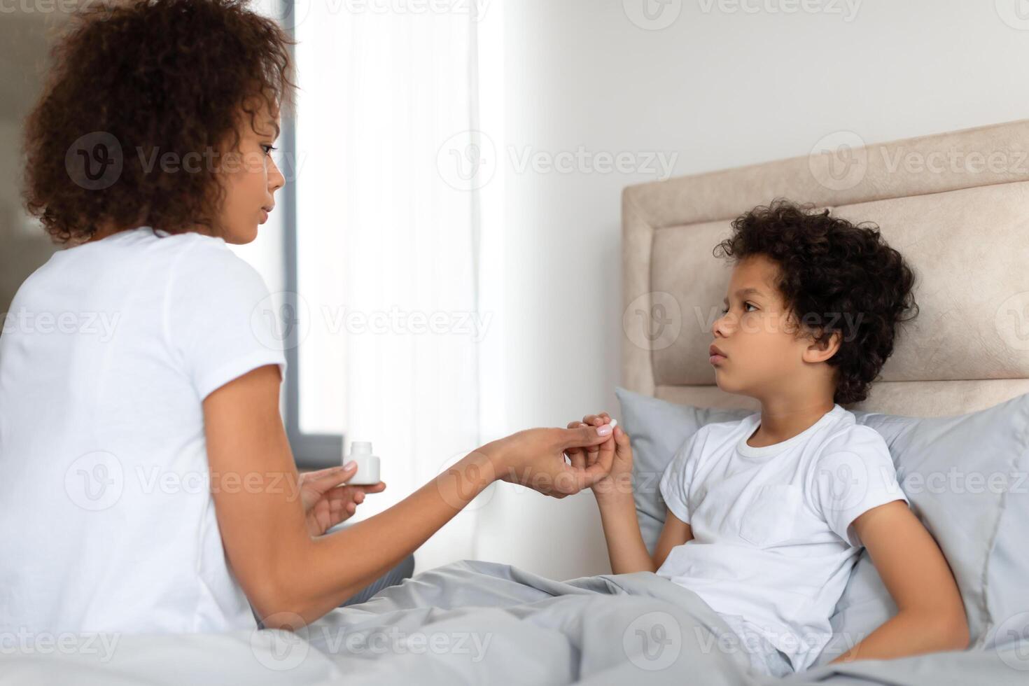 A concerned African American mother sits on the edge of a bed, gently holding her young child hand as she checks for a fever. The child, looking unwell photo