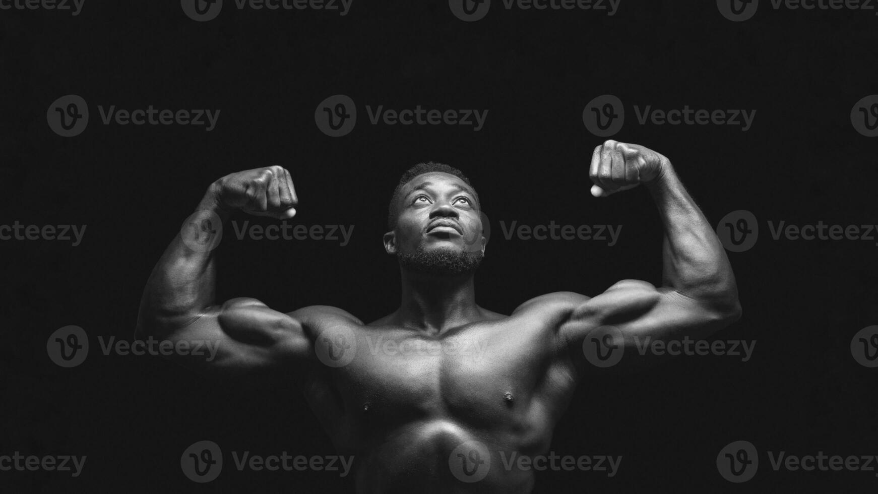 A black and white photo of a man flexing his biceps, looking upward against a dark background.